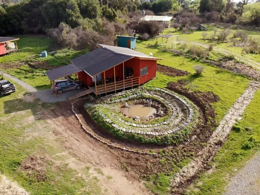 Vista aérea de La Bella Ecoaldea en Rari: casa de madera junto a una laguna en espiral rodeada de bancales en regeneración